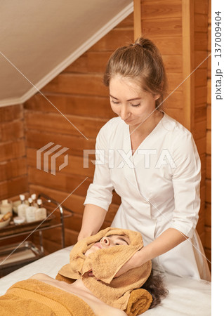 Female Adult Providing Relaxing Spa Treatment To Young Caucasian Woman In Serene Wooden Environment 137009404