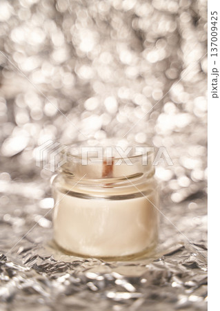 Small glass candle jar with warm glow on shimmering metallic background 137009425