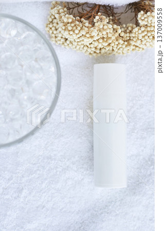 Minimalist skincare setup with white bottle on soft towel surrounded by flowers and glass beads 137009558
