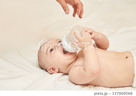 Caucasian Baby Drinking Milk From Bottle On Bed With Adult Hand Assisting Caucasian Baby Drinking Milk From Bottle On Bed With Adult Hand Assisting 137009566