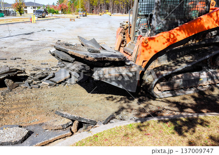 Skid steer machinery works to break up remove old asphalt at construction site in residential neighborhood. Skid steer machinery works to break up remove old asphalt at construction site in residential neighborhood. 137009747