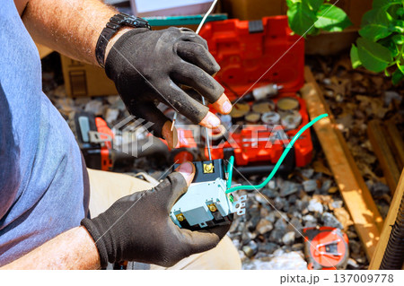 Worker connects wires to device while using tools in workshop area filled with supplies Worker connects wires to device while using tools in workshop area filled with supplies 137009778