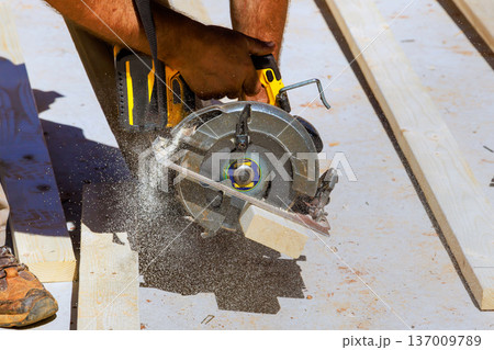 Worker uses circular saw to cut wood at construction site dust flies as blade moves through boards. 137009789