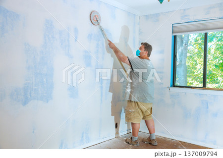 Contractor is sanding wall of room with roller sender while wearing mask for safety. 137009794