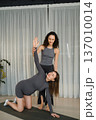 Yoga instructor helps student with stretch in studio during morning class with natural light and curtains 137010014