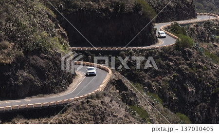 Tenerife, Canary Islands: White cars driving winding mountain road with concrete barriers under a sunny sky, highlighting island's stunning rocky landscape. Transportation, travel. Drone flight 137010405