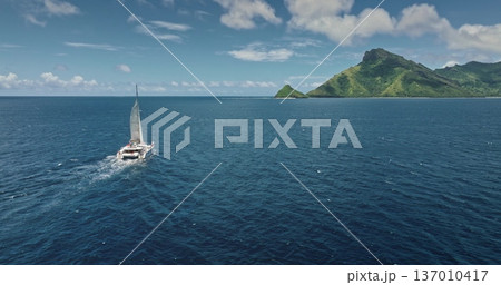 Fiji: Catamaran sailing blue ocean near tropical island with lush vegetation and turquoise water, perfect travel destination for summer vacation. Remote wild nature paradise. Drone flight 137010417