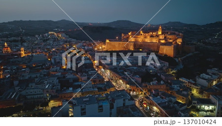Malta, Comino island: Cittadella, historic fortified city, illuminating Victoria's skyline during twilight. Road traffic, streets, church and buildings glowing against dark sky. Aerial flight 137010424