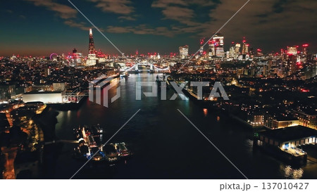 England, London: Breathtaking aerial view London's illuminated skyline at night, Tower Bridge and The Shard reflecting on River Thames. Popular tourist attraction and iconic landmark. Rise up panorama England, London: Breathtaking aerial view London's illuminated skyline at night, Tower Bridge and The Shard reflecting on River Thames. Popular tourist attraction and iconic landmark. Rise up panorama 137010427