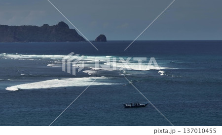 Lombok: Pantai Tomang Omang shoreline with a long white waves break of surf across deep blue sea, rugged island coastline in background and a small fishing boat on tranquil waters. Aerial drone flight Lombok: Pantai Tomang Omang shoreline with a long white waves break of surf across deep blue sea, rugged island coastline in background and a small fishing boat on tranquil waters. Aerial drone flight 137010455
