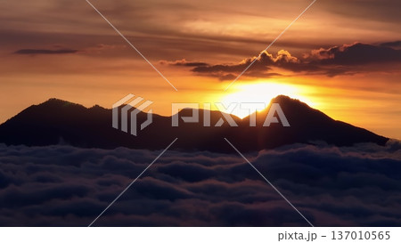 Mount Batur volcano silhouette at sunrise, with golden sunrays peaking over the mountain peak, creating a dramatic skyscape above a thick blanket of clouds. Travel to Bali, Indonesia. Aerial panorama 137010565