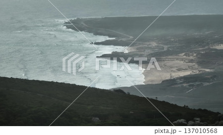 Portugal, Lisbon: Aerial capturing the rugged coastline of Sintra Cascais Natural Park, revealing powerful ocean waves crashing onto sandy beach and rocky shores under a misty, stormy sky 137010568