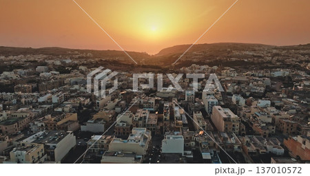 Malta, Comino island: Stunning aerial view city landscape capturing a vibrant sunset over a densely packed townscape. Street buildings under bright orange sky. Drone flight 137010572