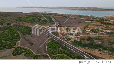 Malta: Cars and bus driving on a road crossing the countryside of Malta island, near the mediterranean sea, with Comino island visible in the background, aerial drone 137010573