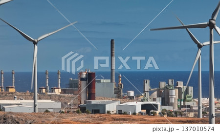 Tenerife, Canary Islands: Spinning wind turbines generating clean energy alongside a factory with smoke-emitting chimneys, highlighting Tenerife's energy transition. Drone flight Tenerife, Canary Islands: Spinning wind turbines generating clean energy alongside a factory with smoke-emitting chimneys, highlighting Tenerife's energy transition. Drone flight 137010574