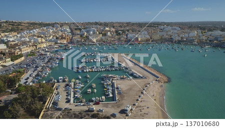 Malta: Aerial view capturing Marsaxlokk harbor in Malta coastline, showcasing colorful boats against backdrop of turquoise Mediterranean water, pier and buildings under blue sky. Drone flight Malta: Aerial view capturing Marsaxlokk harbor in Malta coastline, showcasing colorful boats against backdrop of turquoise Mediterranean water, pier and buildings under blue sky. Drone flight 137010680