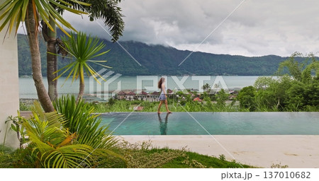 Indonesia, Bali: Woman enjoying tropical vacation, walking along edge of an infinity pool with stunning views of Lake Batur and surrounding green mountains in cloudy day. Relax, summer travel concept Indonesia, Bali: Woman enjoying tropical vacation, walking along edge of an infinity pool with stunning views of Lake Batur and surrounding green mountains in cloudy day. Relax, summer travel concept 137010682