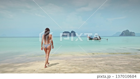 Thailand, Koh Lanta: Woman walking barefoot on sand tropical beach to turquoise water and traditional Thai longtail boat, distant limestone islands framing a tranquil vacation travel escape. Back view 137010684