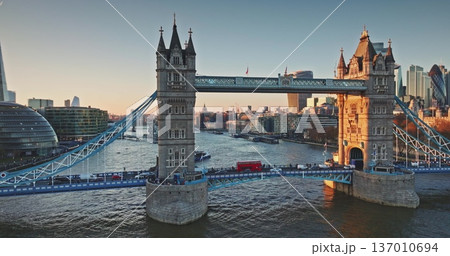Tower Bridge Victorian Gothic towers and bustling cars traffic cross the River Thames at golden hour sunset, London skyline glowing in warm evening light. Travel destination. Aerial panorama footage 137010694