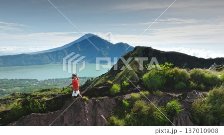 Bali, Indonesia: Caucasian young tourist man enjoying hike along the rugged Batur volcano crater rim, Lake Batur and Mount Agung under bright sky. Travel destination, wild nature landscape scene 137010908