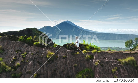 Man running along a mountain ridge above Lake Batur with Mount Agung in background, drone aerial view capturing adventure, sport, freedom and active outdoor exploration in Bali tropical landscape Man running along a mountain ridge above Lake Batur with Mount Agung in background, drone aerial view capturing adventure, sport, freedom and active outdoor exploration in Bali tropical landscape 137010914