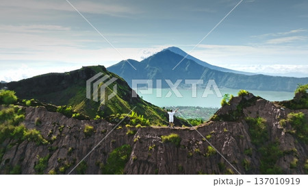 Man celebrating success on mountain ridge with outstretched arms, enjoying panoramic view of caldera, Lake Batur, and Mount Agung in the distance, representing achievement and freedom. Aerial flight Man celebrating success on mountain ridge with outstretched arms, enjoying panoramic view of caldera, Lake Batur, and Mount Agung in the distance, representing achievement and freedom. Aerial flight 137010919