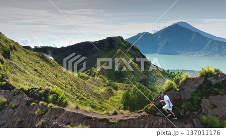 Indonesia, Bali: Man running narrow trail on green crater rim of Mount Batur with an active steam vent, Lake Batur and distant mountains. Sport, hiking, active lifestyle. Aerial view zoom out panorama 137011176