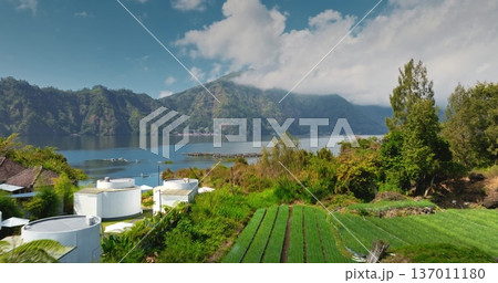 Mount Abang and Lake Batur with agricultural fields in Bali. Lush green terraced agriculture with traditional houses, tropical forest forming a serene landscape in Bali, Indonesia. Aerial view flight Mount Abang and Lake Batur with agricultural fields in Bali. Lush green terraced agriculture with traditional houses, tropical forest forming a serene landscape in Bali, Indonesia. Aerial view flight 137011180