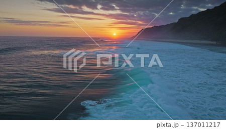 Golden sunset over Indian Ocean coastline, bright colorful clouds sky reflects on vast water surface, powerful waves break tropical Bali island coast with lush green cliffs in background. Aerial shot Golden sunset over Indian Ocean coastline, bright colorful clouds sky reflects on vast water surface, powerful waves break tropical Bali island coast with lush green cliffs in background. Aerial shot 137011217