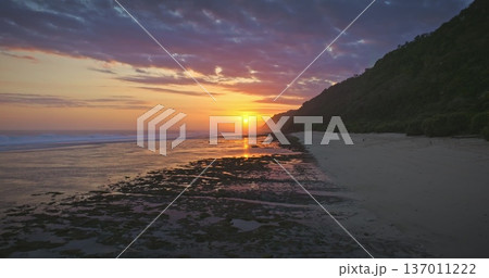 Indonesia, Bali: beautiful colorful sunset painting sky with warm colors, reflecting on low tide ocean beach with exposed reef formations and green lush coastline in background. Aerial drone flight Indonesia, Bali: beautiful colorful sunset painting sky with warm colors, reflecting on low tide ocean beach with exposed reef formations and green lush coastline in background. Aerial drone flight 137011222