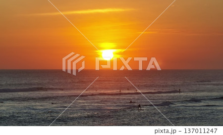 Indonesia, Bali: surfers paddle and ride boards on peaceful ocean waves at bright orange sunset, creating a tranquil scene with sun reflecting golden light on the water and horizon. Natural background 137011247
