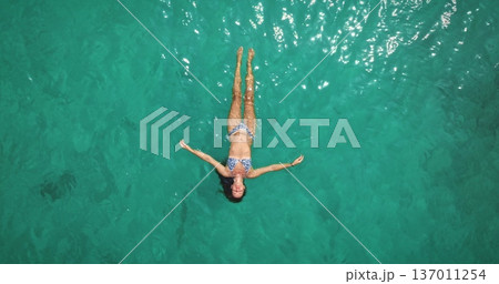 Beautiful young woman relax and enjoy warm, clear sea turquoise water, floating effortlessly on her back, feeling freedom and tranquility during a tropical summer vacation. Aerial top down view Beautiful young woman relax and enjoy warm, clear sea turquoise water, floating effortlessly on her back, feeling freedom and tranquility during a tropical summer vacation. Aerial top down view 137011254
