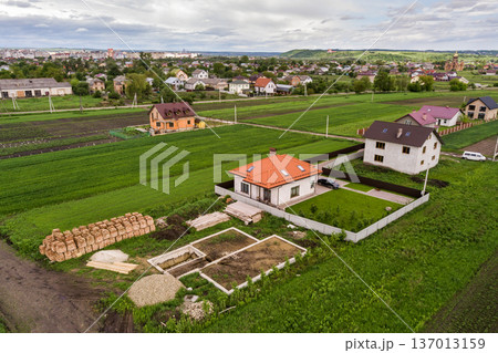 Aerial top view of a private house with attic windows on roof, paved yard with green grass lawn and building site with concrete foundation floor and stacks of yellow bricks for construction.. Aerial top view of a private house with attic windows on roof, paved yard with green grass lawn and building site with concrete foundation floor and stacks of yellow bricks for construction.. 137013159