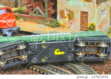 Model train car derailed on tracks near a miniature village scene with a water wheel. Close-up view of a green model train car overturned on a track, with a red locomotive and village buildings in the Model train car derailed on tracks near a miniature village scene with a water wheel. Close-up view of a green model train car overturned on a track, with a red locomotive and village buildings in the 137013454