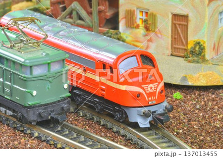 Close-up of two model trains, one red and one green, on miniature railway tracks. Detailed view of a vintage model train set featuring a red M V locomotive alongside a green electric engine on a 137013455