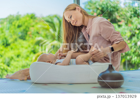 Newborn baby resting in a cozy cocoon beside mother with aroma lamp, calm home atmosphere. Infant relaxation, gentle care, motherhood routine, aromatherapy for babies and family wellbeing concept. 137013929