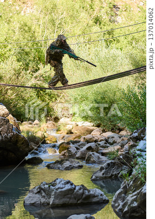 Camouflaged Sniper Soldier in Ghillie Suit Crossing a Narrow Rope Bridge Over Forest Stream 137014062