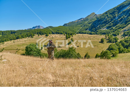 Camouflaged Sniper Standing In Mountain Valley Overlooking Grassy Hills And Forested Slopes 137014083