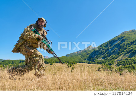 Soldier in Camouflage Ghillie Suit With Rifle on Mountain Surveillance in Sunny Field 137014124
