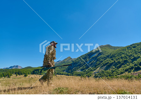 Soldier In Ghillie Suit With Sniper Rifle Walking Across Mountain Meadow Under Blue Sky 137014131