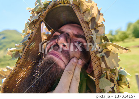 Camouflaged Soldier With Face Paint and Ghillie Hood Preparing in Outdoor Field Camouflaged Soldier With Face Paint and Ghillie Hood Preparing in Outdoor Field 137014157