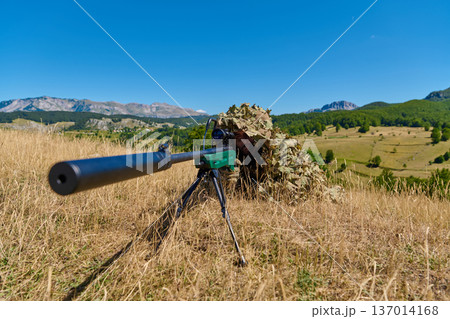 Sniper In Ghillie Suit Aiming A Rifle With Suppressor In Open Grassland Landscape Sniper In Ghillie Suit Aiming A Rifle With Suppressor In Open Grassland Landscape 137014168