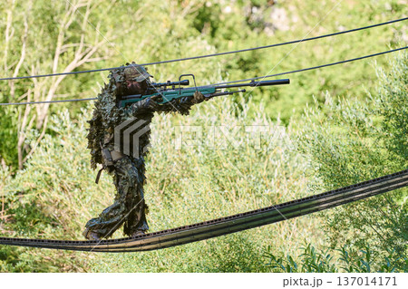 Sniper Soldier in Ghillie Suit Aiming Rifle While Crossing Suspended Rope Bridge During Military Recon Training Sniper Soldier in Ghillie Suit Aiming Rifle While Crossing Suspended Rope Bridge During Military Recon Training 137014171