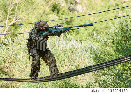 Sniper Soldier in Ghillie Camouflage Aiming Rifle While Crossing Suspended Cable Bridge in Woodland 137014173