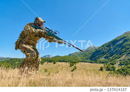 Sniper Soldier In Ghillie Camouflage Aiming Precision Rifle On Mountain Ridge Over Valley 137014187