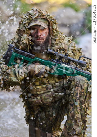 Camouflaged Sniper Soldier In Ghillie Suit With Scoped Rifle Kneeling In Woodland Stream Camouflaged Sniper Soldier In Ghillie Suit With Scoped Rifle Kneeling In Woodland Stream 137014215