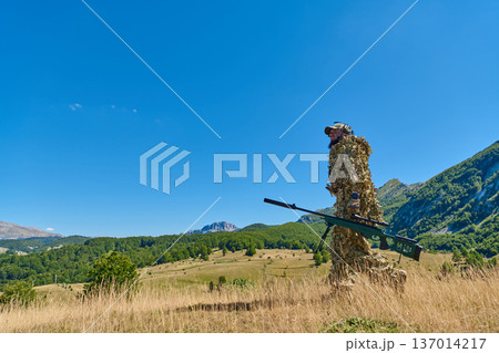 Camouflaged Sniper Soldier In Ghillie Suit Carrying Sniper Rifle Across Mountain Meadow Landscape 137014217