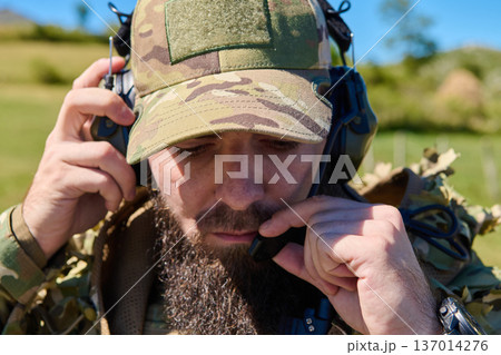 Military Radio Operator in Camouflage Gear Wearing Headset and Microphone During Field Operation Military Radio Operator in Camouflage Gear Wearing Headset and Microphone During Field Operation 137014276