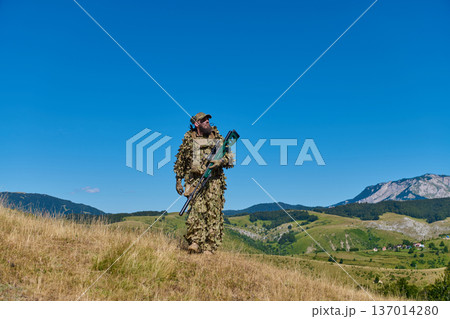 Soldier In Ghillie Suit With Sniper Rifle Patrolling Mountain Grassland Under Clear Blue Sky 137014280