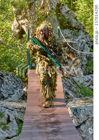 Bearded Soldier In Ghillie Suit Crossing Forest Bridge With Sniper Rifle During Sunny Recon Mission Bearded Soldier In Ghillie Suit Crossing Forest Bridge With Sniper Rifle During Sunny Recon Mission 137014290